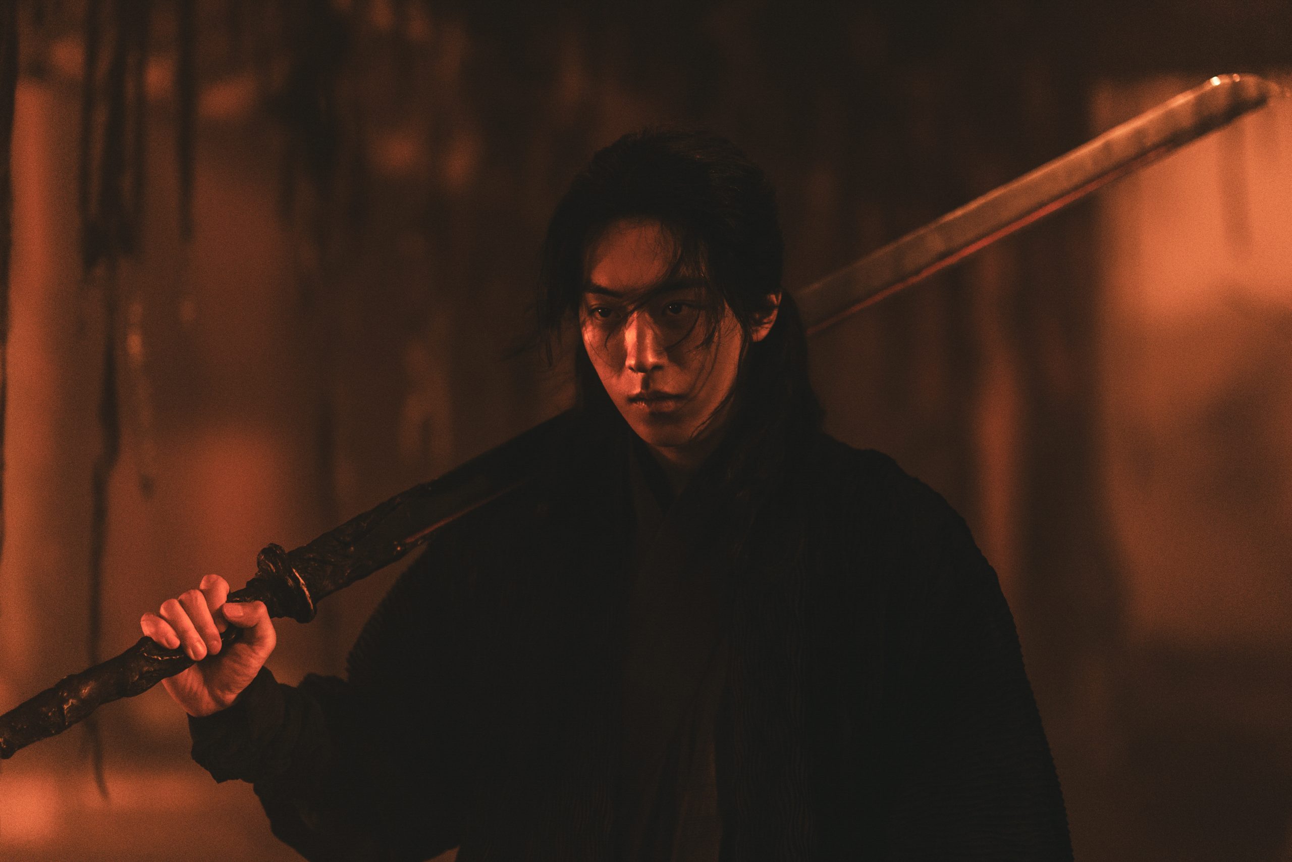 "A person dressed in dark clothing stands in a dimly lit, smoky environment, holding a large sword over their shoulder, with a serious and intense expression on their face."